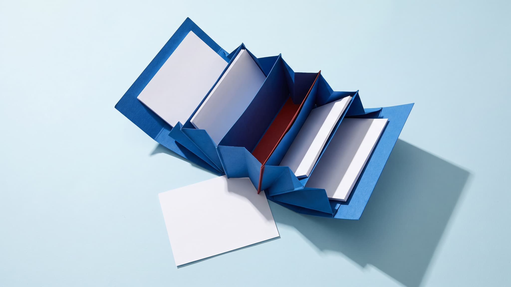 Top-down view of a cobalt blue accordion document folder on an ice-blue surface with blank white sheets and one empty slot marked by a vermilion accent